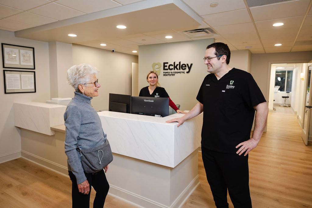 Dr. Ryan Eckley and a team member greet a smiling senior patient at the reception desk of Eckley Family and Cosmetic Dentistry in Wilmington, DE.